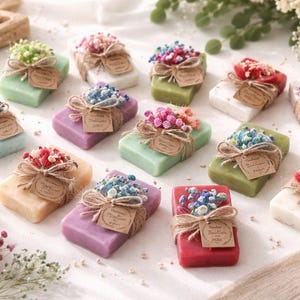 May include: A selection of colourful soaps, each wrapped with twine and decorated with dried flowers. Soaps are various colours, including green, purple, and white. Each soap has a small tag attached.