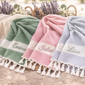 May include: A display of personalized beach towels in various colors: beige, green, pink, and blue. Each towel features a white stripe with a name embroidered on it, and white tassels. Baskets with flowers are in the background.