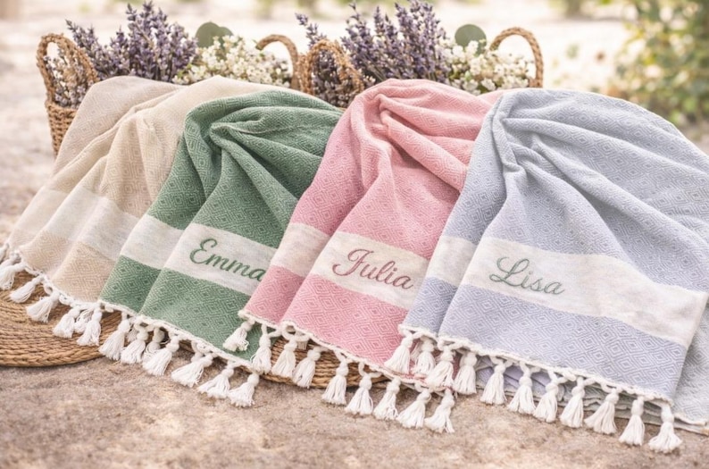 May include: A display of personalized beach towels in various colors: beige, green, pink, and blue. Each towel features a white stripe with a name embroidered on it, and white tassels. Baskets with flowers are in the background.