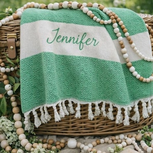 May include: A green and white striped blanket with the name "Jennifer" embroidered on it, resting on a wicker basket. The blanket has a diamond pattern and white fringe. Wooden bead garlands and flowers surround the basket.