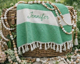 May include: A green and white striped blanket with the name "Jennifer" embroidered on it, resting on a wicker basket. The blanket has a diamond pattern and white fringe. Wooden bead garlands and flowers surround the basket.