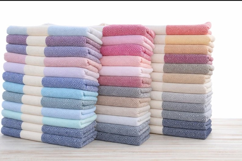 May include: Stacks of colorful, folded Turkish towels. The towels feature a diamond pattern and are in shades of blue, pink, and neutral tones. The towels are stacked neatly, ready for use.