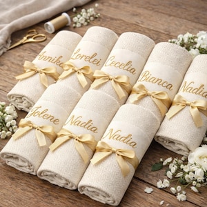May include: Rolled cream-colored towels with gold embroidered names and matching gold ribbon bows. The towels have a geometric pattern and are arranged on a wooden surface, with floral accents and sewing supplies in the background.