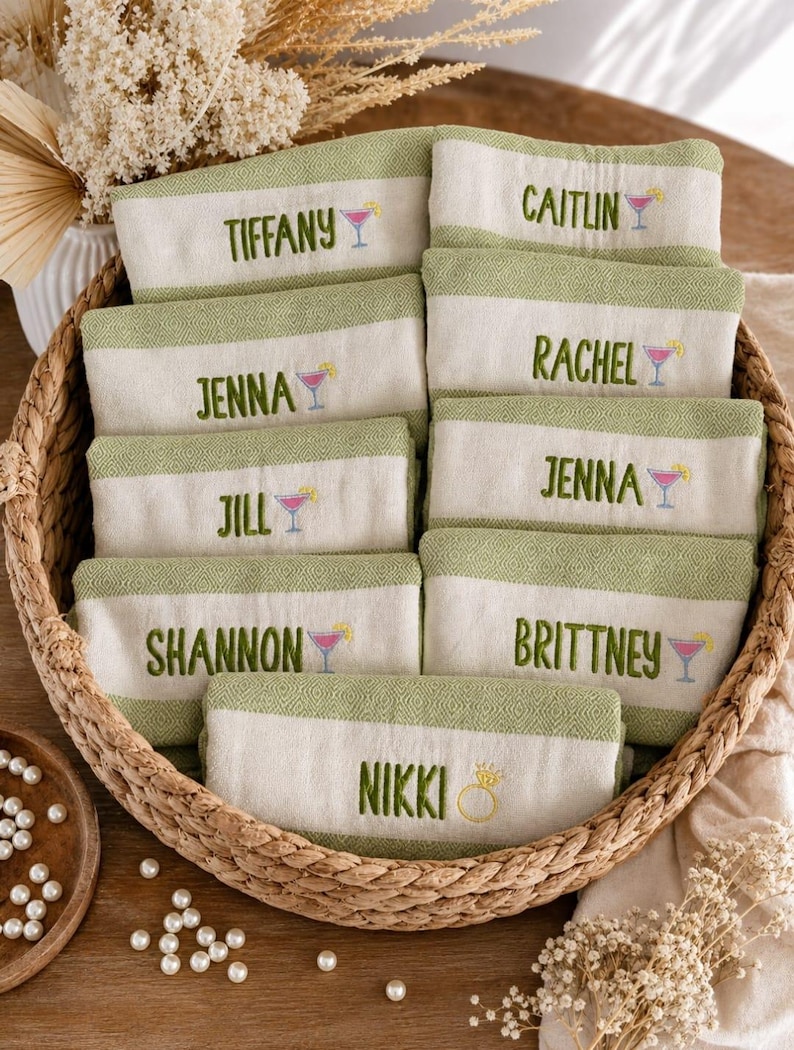 May include: A woven basket filled with personalized hand towels. Each towel is white with a green border and features an embroidered name and a cocktail glass. Names include Tiffany, Caitlin, Jenna, Rachel, Jill, Jenna, Shannon, Brittney, and Nikki.