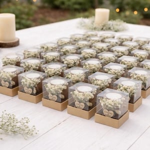 May include: Arrangement of small, square gift boxes with clear tops, each containing a white candle and a small bouquet of white flowers. The boxes are on a white wooden surface, with two larger candles in the background.