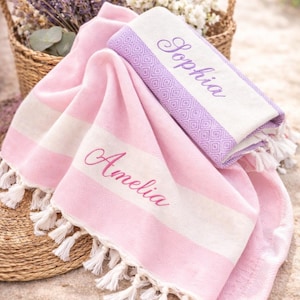May include: Two personalized beach towels in pink and white with the names "Amelia" and "Sophia" embroidered in pink. One towel is folded, and the other is draped. Both have white fringe. A basket of lavender and flowers is in the background.