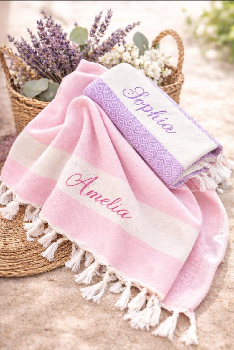 May include: Two personalized beach towels in pink and white with the names "Amelia" and "Sophia" embroidered in pink. One towel is folded, and the other is draped. Both have white fringe. A basket of lavender and flowers is in the background.