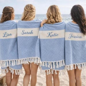 May include: Four people stand on a beach, each draped in a light blue towel with a white stripe. The towels have fringed edges and personalized names embroidered on the white stripe: Lisa, Sarah, Katie, and Jessica. The ocean is in the background.