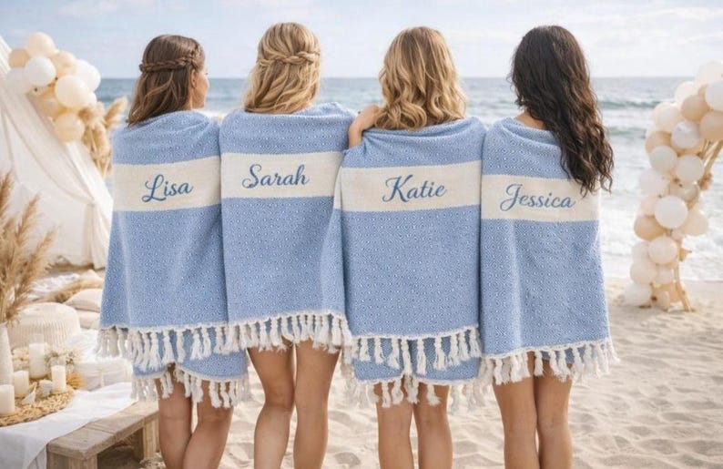 May include: Four people stand on a beach, each draped in a light blue towel with a white stripe. The towels have fringed edges and personalized names embroidered on the white stripe: Lisa, Sarah, Katie, and Jessica. The ocean is in the background.