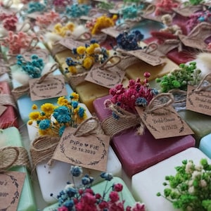 May include: A collection of colourful soaps, each decorated with dried flowers and a small tag tied with twine. The soaps are in various shades, including white, green, purple, and yellow. Some tags read "Pipe FIE 29.10.2020" and "Christina's Bridal Shower 10/25/20".
