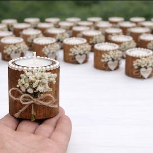 May include: A collection of decorative wooden candle holders. Each holder is a short, cylindrical piece of wood, adorned with a small bouquet of white flowers, a heart-shaped embellishment, and a twine bow. The candle holders are topped with a white candle.