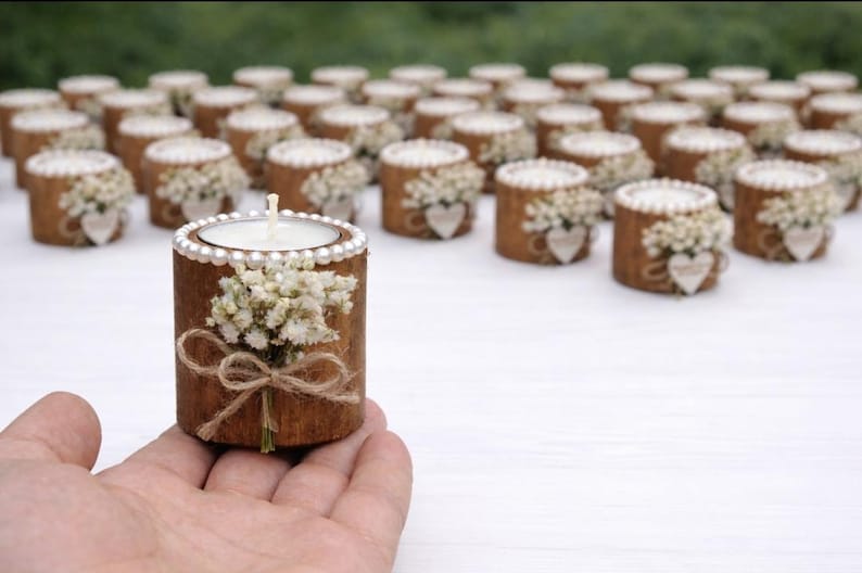 May include: A collection of decorative wooden candle holders. Each holder is a short, cylindrical piece of wood, adorned with a small bouquet of white flowers, a heart-shaped embellishment, and a twine bow. The candle holders are topped with a white candle.