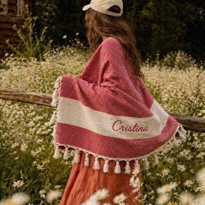 May include: A red and white patterned shawl with white tassels and the name "Cristina" embroidered on it. The shawl is draped over a person in a field of white flowers. A beige cap is worn.