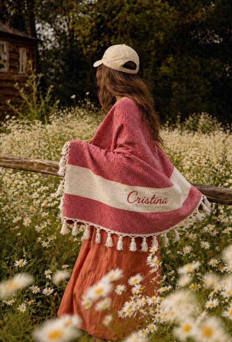 May include: A red and white patterned shawl with white tassels and the name "Cristina" embroidered on it. The shawl is draped over a person in a field of white flowers. A beige cap is worn.