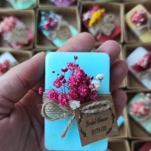 May include: A turquoise soap bar adorned with pink and white dried flowers, secured with twine and a small tag reading "Bridal Shower 10/25/20". The soap is held in a hand, with other similar soaps visible in the background.