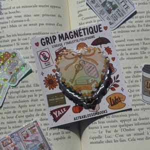 May include: A heart-shaped magnetic grip for e-readers, tablets, and phones on a white backing card. The design features a gold key, books, autumn leaves, and mushrooms. Text includes GRIP MAGNÉTIQUE, STARS HOLLOW, YALE, LUKE'S, and ASTRABLOSSOMBOOKS.