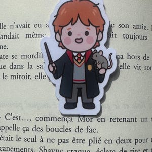 Può includere: Un segnalibro magnetico con un'illustrazione a cartoni animati di una giovane persona dai capelli rossi, che indossa una tunica scura sopra un maglione grigio, cravatta rossa e camicia bianca. Il personaggio tiene una bacchetta nera e porta un piccolo topo grigio.