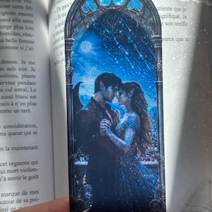 Op de afbeelding: Een rechthoekige boekenlegger met een gotisch spitsboogvenster waarin twee figuren met puntige oren dansen onder een sterrenhemel. De illustratie heeft een donkerblauw kleurenpalet met glinsterende holografische accenten die het geheel oplichten.