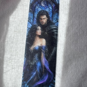 Peut inclure: Marque-page avec une illustration fantastique d'un couple. La femme porte une robe bleue avec des accents violets, et l'homme est en armure sombre. Le fond est un vitrail avec un croissant de lune.