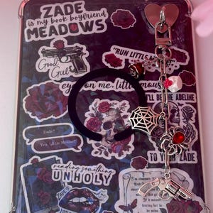 Op de afbeelding: Een zwarte tablethoes versierd met stickers en een bedel. De stickers tonen tekst, rozen, schedels en een pistool. Een zwarte elastische band en een zilveren bedel met een hart, spinnenweb en andere bedels zijn bevestigd.