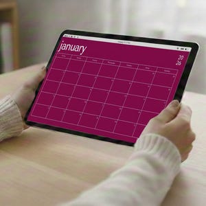 May include: A digital tablet showing a January 2026 calendar. The calendar grid is a deep magenta colour, with the word "January" at the top. The tablet is held by a person wearing a white sweater, with a pen and coffee cup on the table.