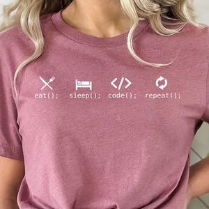May include: A mauve-colored t-shirt with white text and symbols. The text reads "eat(); sleep(); code(); repeat();" with corresponding symbols for each action. The shirt is made of a soft material.