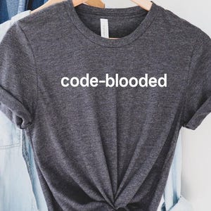 May include: Dark gray t-shirt with the text "code-blooded" in white. The shirt is hanging on a wooden hanger with other clothing items, including a light blue shirt and a brown hat.