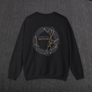 May include: Black sweatshirt featuring a dark gray and gold design on the back. The design is a broken circle with gold lines and the text "Fall seven times Stand up eight". The sweatshirt is a crew neck style.