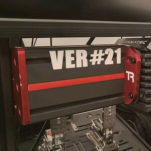 Peut inclure: Configuration de simulateur de course noir et rouge avec un volant et un écran. Le volant comporte de nombreux boutons et un affichage numérique. Le texte "VER #21" est visible sur un composant noir et rouge.