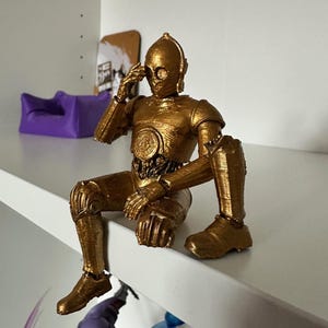 Könnte beinhalten: Goldfarbene C-3PO-Actionfigur aus Star Wars, sitzend auf einem weißen Regal. Die Figur hat bewegliche Gelenke und ein metallisches Finish. Die Figur ist mit einer Hand am Kopf posiert.