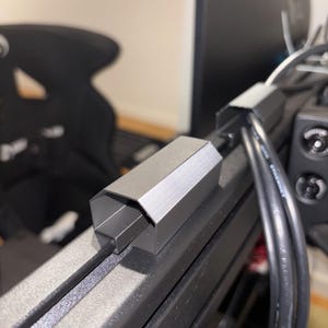 Può includere: Una clip geometrica argentata per la gestione dei cavi è attaccata a un telaio metallico nero. Sono visibili diversi cavi neri, insieme a un monitor del computer e a una sedia nera sullo sfondo. La clip sembra essere in alluminio.