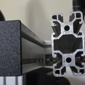 Peut inclure: Gros plan de profils d'extrusion en aluminium, montrant une section transversale et un joint d'angle. Les profils sont argentés et noirs, avec un embout noir texturé. La conception suggère une application structurelle ou mécanique.