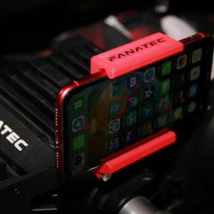 Peut inclure: Un smartphone rouge est maintenu dans un support rouge vif, avec le mot "FANATEC" imprimé sur le support. Le téléphone affiche un écran d'icônes d'applications. Le support est monté sur un appareil noir, peut-être un accessoire de jeu.