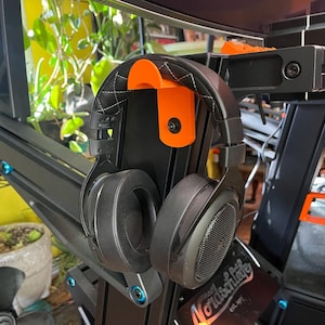 Op de afbeelding: Zwarte over-ear hoofdtelefoon hangt aan een oranje en zwarte hoofdtelefoonhouder. De houder is bevestigd aan een zwart metalen frame. De hoofdtelefoon heeft een zwarte gewatteerde hoofdband en grote oorschelpen. Het frame heeft blauwe accenten.