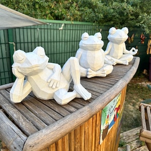 Puede incluir: Tres figuras de ranas geométricas de color blanco roto dispuestas sobre una superficie de madera. Una rana está recostada de lado, mientras que las otras dos están sentadas en posturas meditativas con los ojos cerrados y facetas.