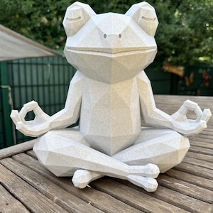 Peut inclure: Une figurine de grenouille géométrique à facettes, de couleur gris clair moucheté, en position de méditation assise. La grenouille a les yeux fermés, un museau pointu et les bras étendus avec les doigts en mudra, présentant une texture angulaire.