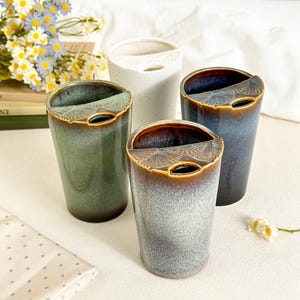 May include: Three ceramic tumblers with a unique ginkgo leaf-shaped rim detail. One tumbler is speckled white, one is a gradient of green to brown, and one is a gradient of blue to brown. Each vessel features a smooth, glazed finish and a cylindrical shape.