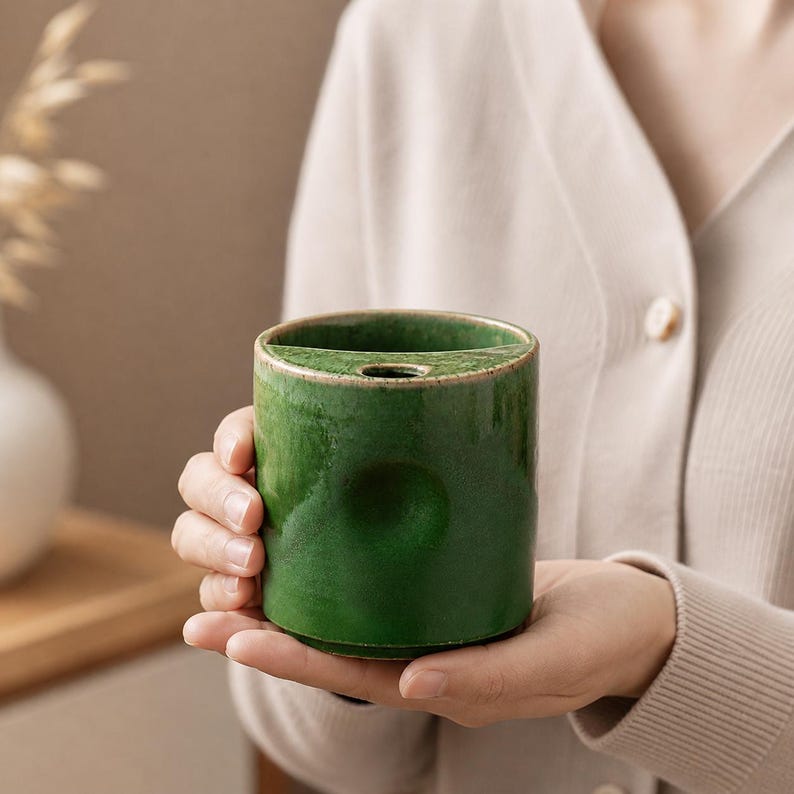 May include: A cylindrical, emerald green ceramic vase with a gold rim. The vase has a unique design with a slot and a small hole, likely for arranging flowers. The glossy finish reflects light, highlighting the rich color and texture of the ceramic.