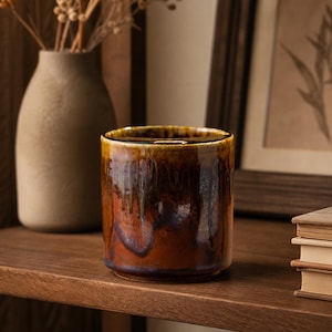 May include: A ceramic, cylindrical vase with a glossy, glazed finish. The vase features a blend of brown and amber tones with a dripped glaze effect. It sits on a wooden shelf, with a neutral-toned vase and framed artwork in the background.