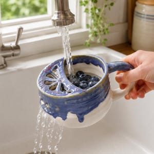 Pode incluir: Uma caneca coador de cerâmica para bagas com topo esmaltado em azul e base branca, apresentando uma seção de coador integrada com um padrão de recortes em forma de flor. A caneca é segurada sob uma torneira, enxaguando mirtilos frescos.