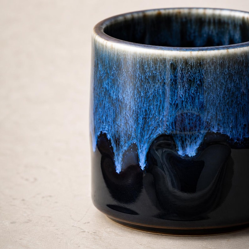 May include: A ceramic mug with a dark blue glaze that drips down the sides, creating a textured effect. The mug has a glossy finish and a contrasting white rim. The bottom of the mug is a dark brown color.