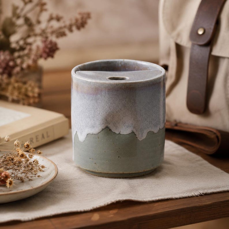 May include: A cylindrical ceramic toothbrush holder with a two-tone glaze. The top half is a light gray with a subtle iridescent sheen, and the bottom half is a muted green. The holder has a single, oval opening at the top.