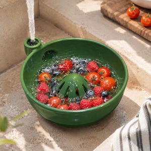 May include: A dark green ceramic berry colander bowl with a central drainage cone and a side water intake spout. The bowl is filled with water, red cherry tomatoes, raspberries, and blueberries, with a stream of water flowing into the spout.
