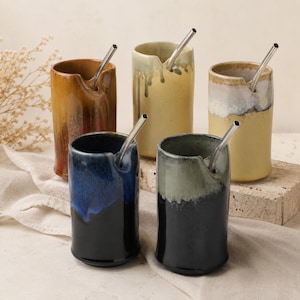 Op de afbeelding: Vijf keramische bekers in verschillende kleuren, waaronder bruin, blauw en groen, elk met een roestvrijstalen rietje. De bekers hebben een uniek ontwerp met een schenktuit. De achtergrond is neutraal, waardoor het kleurrijke drinkgerei opvalt.