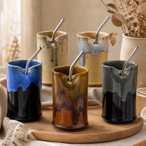 May include: A set of five ceramic tumblers with stainless steel straws. Each tumbler features a unique color scheme, including blue, brown, and gray, with a dripped glaze effect. The tumblers are displayed on a wooden tray, showcasing their handcrafted appearance.