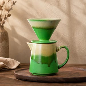 Może przedstawiać: Zestaw ceramicznego ekspresu do kawy z przelewem. Zestaw zawiera zielono-kremowy dripper, pasującą pokrywkę oraz zielono-kremową karafkę z uchwytem. Zestaw znajduje się na drewnianej tacy.