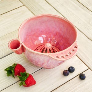 Op de afbeelding: Een roze keramische citruspers met een glanzende afwerking, voorzien van een centrale perskegel, een handvat aan de zijkant met vijf kleine ronde gaatjes en een kleine tuit aan de tegenoverliggende zijde. Twee verse rode aardbeien en twee blauwe bessen liggen ernaast.