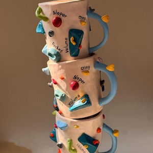 Puede incluir: Tres tazas de cerámica apiladas con asas azul claro y diseños de presas de escalada. Cada taza presenta presas de escalada coloridas y texturizadas en varias formas y las palabras "sloper", "crimp", "pocket", "volume" y "jug".