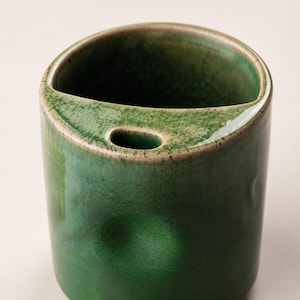 May include: A cylindrical, green ceramic vase with a glossy finish. The vase has a unique, curved opening at the top and a small, oval hole on the side. The design is simple and modern, suitable for holding small items or as a decorative piece.