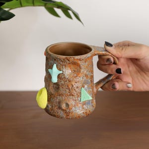 May include: A handmade ceramic mug with a textured, mottled brown and orange surface. The mug features a handle and is adorned with felt shapes in yellow, light blue, and green. The mug is held by a person.